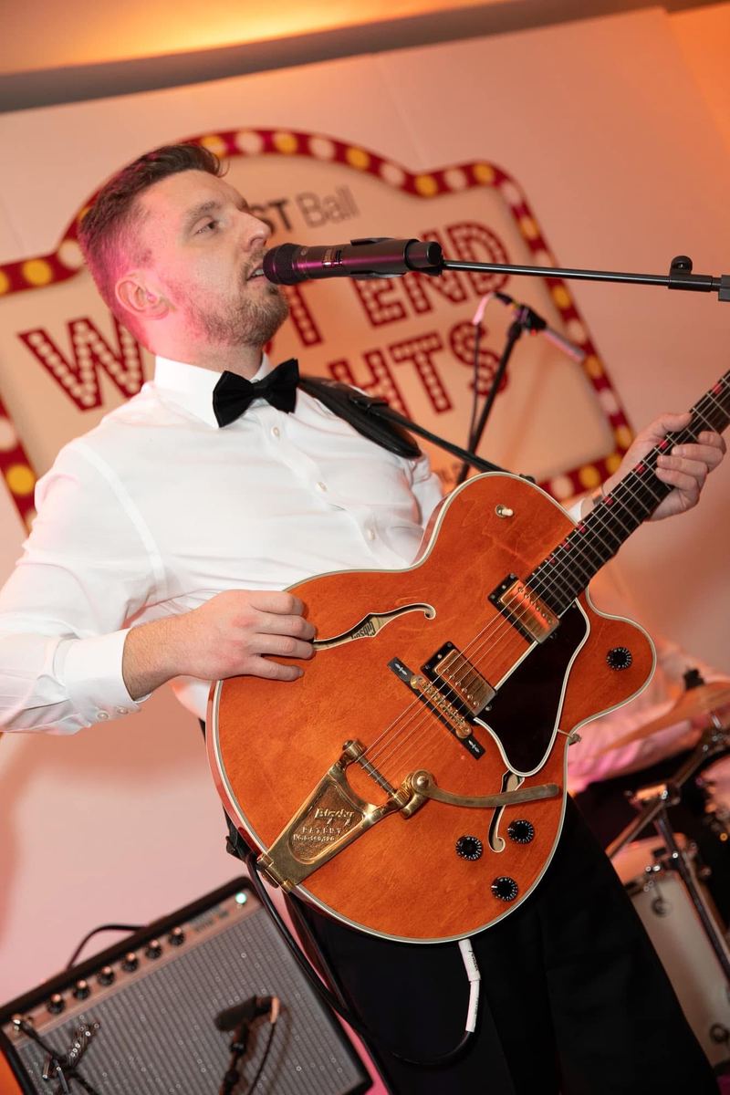 Tom in a bow tie singing and playing an orange hollow-body guitar