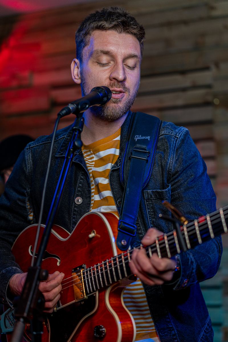 Tom singing and playing a red hollow-body guitar in a denim jacket