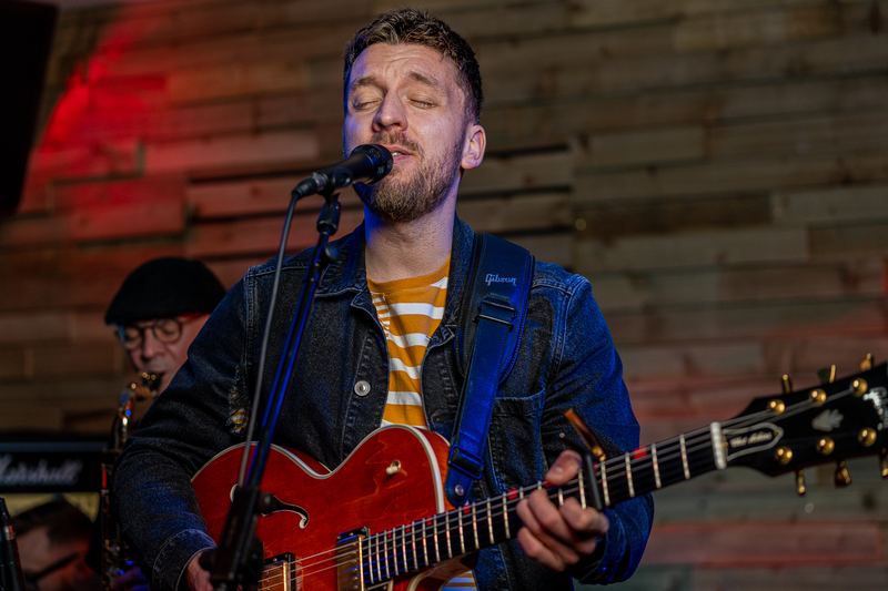 Tom singing into a microphone with a red guitar, saxophonist behind